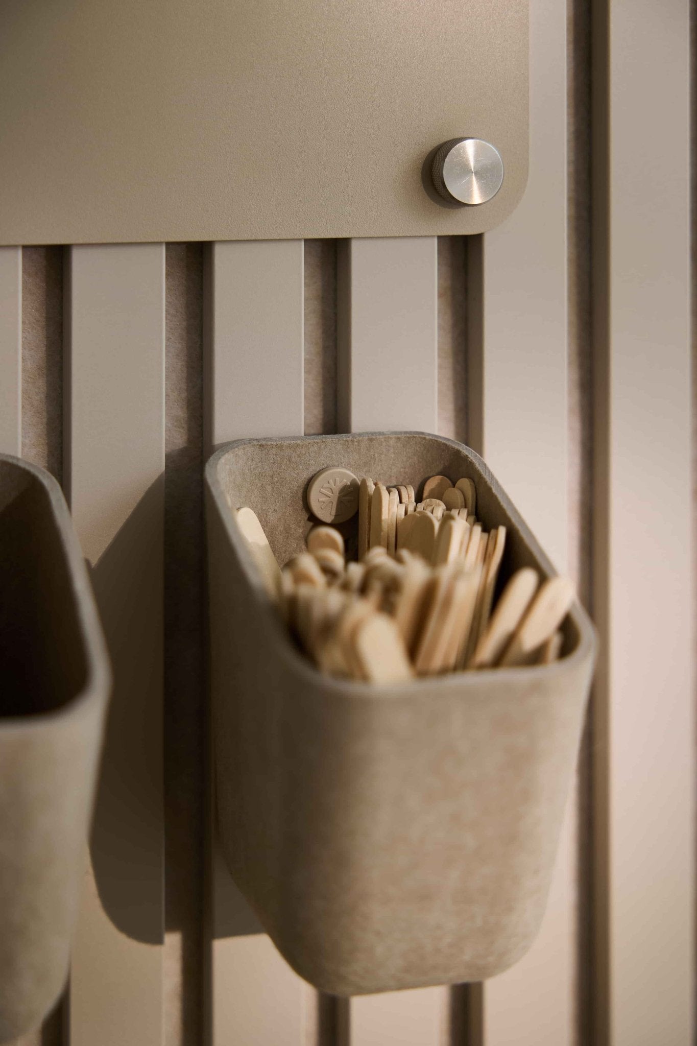 WoodUpp Dupps | En ljus förvaringsenhet i beige för omrörningspinnar och skedar, monterad på en akustikpanel av typen Nordic Akupanel med en metallknopp ovanför för att öppna den.